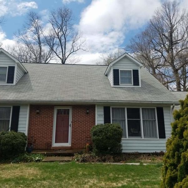 A suburban house with a gray roof and two dormer windows sits under a partly cloudy sky. The lower level, beautifully accentuated with brick, features a welcoming red front door and large inviting windows. Surrounding it are well-maintained bushes and a lush lawn with a charming small tree in the foreground. For residents in Harford County MD, Baltimore County MD, Cecil County MD, Howard County MD, Carroll County MD, Anne Arundel County MD; Wilmington DE; New Castle County DE; or Delaware County PA looking to enhance their home's curb appeal just like this one, Maryland Pro Wash offers expert pressure washing services that can make your home shine and stand out in any neighborhood.