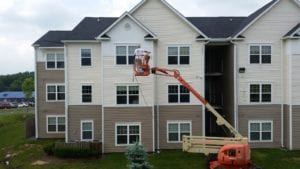 A worker on an orange lift carefully paints the exterior of a charming three-story residential building, which features a classic combination of beige and white siding accented by black shingles. The surrounding area is lush and green, dotted with mature trees and shrubs that enhance the property’s aesthetic appeal. For homeowners looking to maintain or restore their property's pristine condition in this scenic environment, Maryland Pro Wash offers top-notch pressure washing services throughout Harford County MD, Baltimore County MD, Cecil County MD, Howard County MD, Carroll County MD, Anne Arundel County MD; as well as Wilmington DE, New Castle County DE; and Delaware County PA. Their expert team can help refresh surfaces with professional exterior cleaning that complements any new paint job.