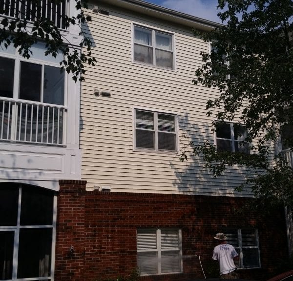 A person wearing a hat stands outside a three-story building with white siding and red brick on the lower level, expertly using a pressure washer to clean the exterior surfaces. Trees partially obscure the view, enhancing the serene setting on this sunny day with a clear sky. This scene is reminiscent of Maryland Pro Wash, a professional business offering top-notch pressure washing services across multiple locations, including Harford County, Baltimore County, Cecil County, Howard County, Carroll County, and Anne Arundel County in Maryland. They also extend their expertise to Wilmington and New Castle County in Delaware as well as Delaware County in Pennsylvania.