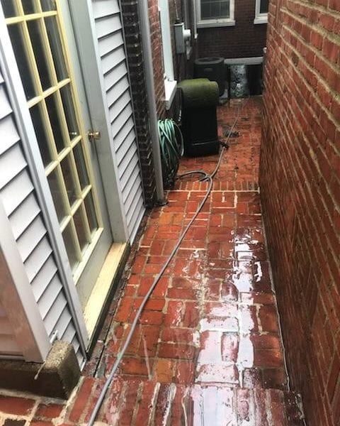 In a narrow brick alleyway adjacent to a building, the wet floor glistens under the light. A coiled garden hose rests on the ground, while a power cord is stretched across the damp area. The glass door leading into the alley reflects the moist surface and is enveloped by vibrant red brick walls. This scene demonstrates how Maryland Pro Wash brings its expert pressure washing services to life, transforming spaces in Harford County MD, Baltimore County MD, Cecil County MD, Howard County MD, Carroll County MD, Anne Arundel County MD within Maryland. Extending their reach beyond state lines into Wilmington DE and New Castle County DE in Delaware as well as Delaware County PA in Pennsylvania. Whether tackling slippery surfaces or maintaining pristine entryways like this one, Marylad Pro wash ensures that every nook and cranny shines with cleanliness.