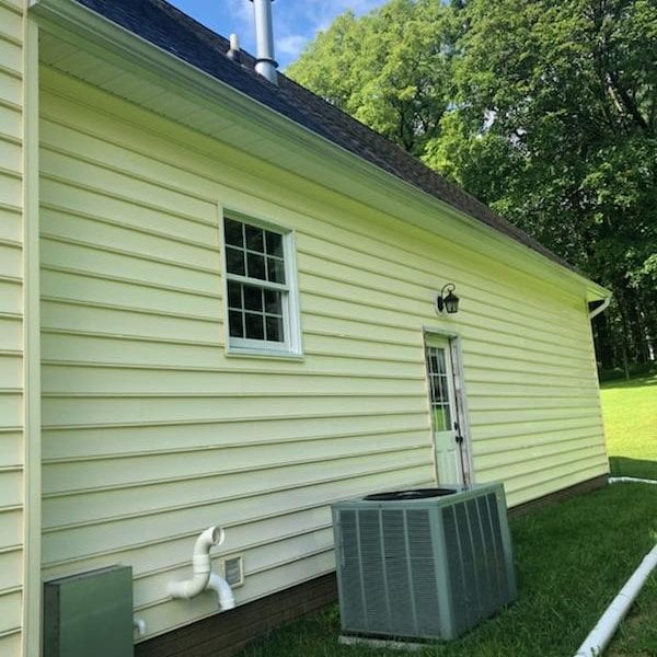 Sure, here is a concise description:

In a serene setting with a blue sky backdrop, imagine the side view of a charming light yellow house. It features a small window and a welcoming door adorned with an overhead light fixture. Adjacent to the door lies an air conditioning unit, enhancing the home's comfort. The house is complemented by a well-maintained grassy lawn and shaded by surrounding trees.

This picturesque scene perfectly illustrates the type of property that Maryland Pro Wash specializes in rejuvenating through their expert pressure washing services. Serving areas like Harford County MD, Baltimore County MD, Cecil County MD, Howard County MD, Carroll County MD, Anne Arundel County MD; as well as Wilmington DE and New Castle County DE; extending up to Delaware County PA – Maryland Pro Wash ensures every element from siding to pathways gleams as fresh and inviting as this idyllic home setting suggests.