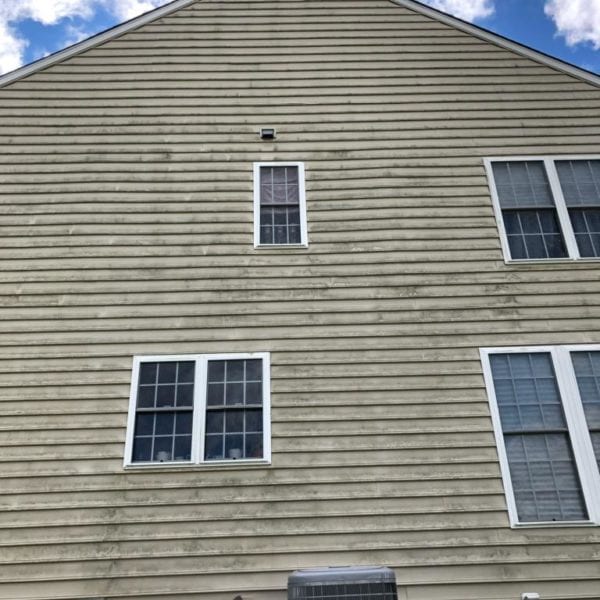 The image depicts the side of a beige house featuring three white-framed windows, set against a backdrop of a blue sky with scattered clouds. Near the base of the house is an air conditioning unit. This scene excellently represents typical residential properties that Maryland Pro Wash services. Specializing in pressure washing, Maryland Pro Wash serves areas including Harford County, Baltimore County, Cecil County, Howard County, Carroll County, and Anne Arundel County in Maryland. Their expertise extends to Wilmington and New Castle County in Delaware, as well as Delaware County in Pennsylvania. Whether it’s removing grime or enhancing curb appeal, they are equipped to help maintain homes just like this one across these regions.