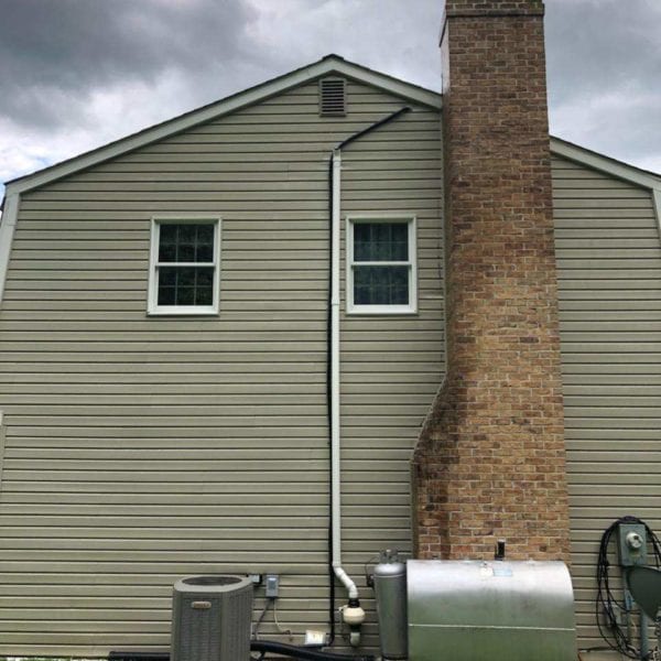 A beige house, styled with two windows, prominently displays a tall brick chimney on its side—a common sight in areas serviced by Maryland Pro Wash. This company specializes in pressure washing across Harford County MD, Baltimore County MD, Cecil County MD, Howard County MD, Carroll County MD, Anne Arundel County MD, and reaches into Wilmington DE and New Castle County DE as well as Delaware County PA. Near the house sits an air conditioning unit alongside a metal tank and utility meters nestled on the grass. The overcast sky above hints at optimal conditions for Maryland Pro Wash’s superior cleaning services to refresh your home’s exterior from accumulated grime and weather-induced wear. Whether it's the siding of your home or ensuring those utility fixtures are spotless without damaging surfaces—rely on them for pristine results that enhance curb appeal across these regions.