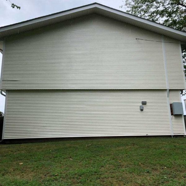 The image depicts a beige, two-story house with horizontal siding, showcasing its clean and well-maintained exterior. This side view features a windowless facade set against a backdrop of lush grass and distant trees beneath a cloudy sky. If your property in Harford County, Baltimore County, Cecil County, Howard County, Carroll County, Anne Arundel County in Maryland or Wilmington and New Castle County in Delaware, or Delaware County in Pennsylvania requires expert pressure washing services to achieve such pristine results, Maryland Pro Wash is here to help. Specializing in comprehensive pressure washing solutions tailored for each unique facet of your home’s exterior needs across these regions.