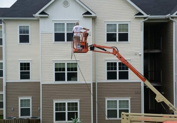 A worker on an orange lift carefully paints the exterior of a charming three-story residential building, which features a classic combination of beige and white siding accented by black shingles. The surrounding area is lush and green, dotted with mature trees and shrubs that enhance the property’s aesthetic appeal. For homeowners looking to maintain or restore their property's pristine condition in this scenic environment, Maryland Pro Wash offers top-notch pressure washing services throughout Harford County MD, Baltimore County MD, Cecil County MD, Howard County MD, Carroll County MD, Anne Arundel County MD; as well as Wilmington DE, New Castle County DE; and Delaware County PA. Their expert team can help refresh surfaces with professional exterior cleaning that complements any new paint job.