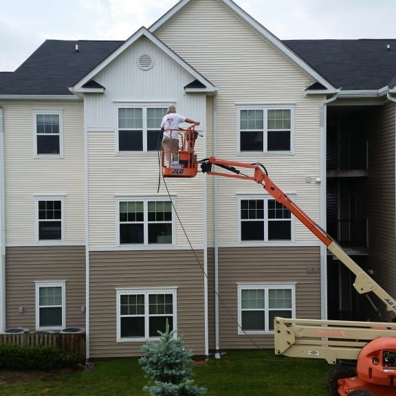 Apartment Complex Pressure Washing | Maryland Pro Wash | Professional Pressure Washing in MD, DE, & PA A worker on an orange lift carefully paints the exterior of a charming three-story residential building, which features a classic combination of beige and white siding accented by black shingles. The surrounding area is lush and green, dotted with mature trees and shrubs that enhance the property’s aesthetic appeal. For homeowners looking to maintain or restore their property's pristine condition in this scenic environment, Maryland Pro Wash offers top-notch pressure washing services throughout Harford County MD, Baltimore County MD, Cecil County MD, Howard County MD, Carroll County MD, Anne Arundel County MD; as well as Wilmington DE, New Castle County DE; and Delaware County PA. Their expert team can help refresh surfaces with professional exterior cleaning that complements any new paint job.