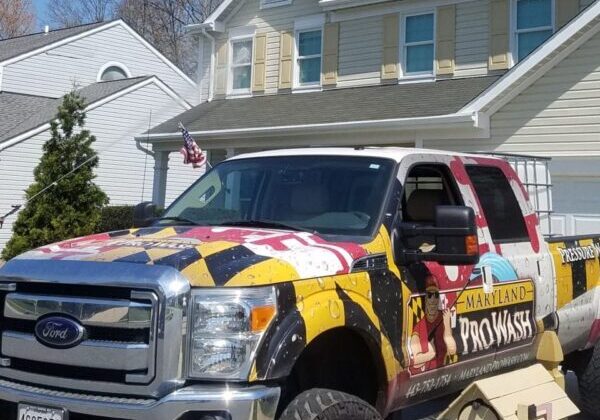 A vibrantly designed ProWash Ford truck, adorned with the distinctive Maryland flag and company branding, is parked in a suburban driveway. This eye-catching vehicle represents Maryland Pro Wash, a premier pressure washing service operating in Harford County MD, Baltimore County MD, Cecil County MD, Howard County MD, Carroll County MD, Anne Arundel County MD, Wilmington DE, New Castle County DE, and Delaware County PA. In the background stands a person positioned in front of one house from a row of similar suburban homes—all potential beneficiaries of Maryland Pro Wash’s top-quality cleaning services.